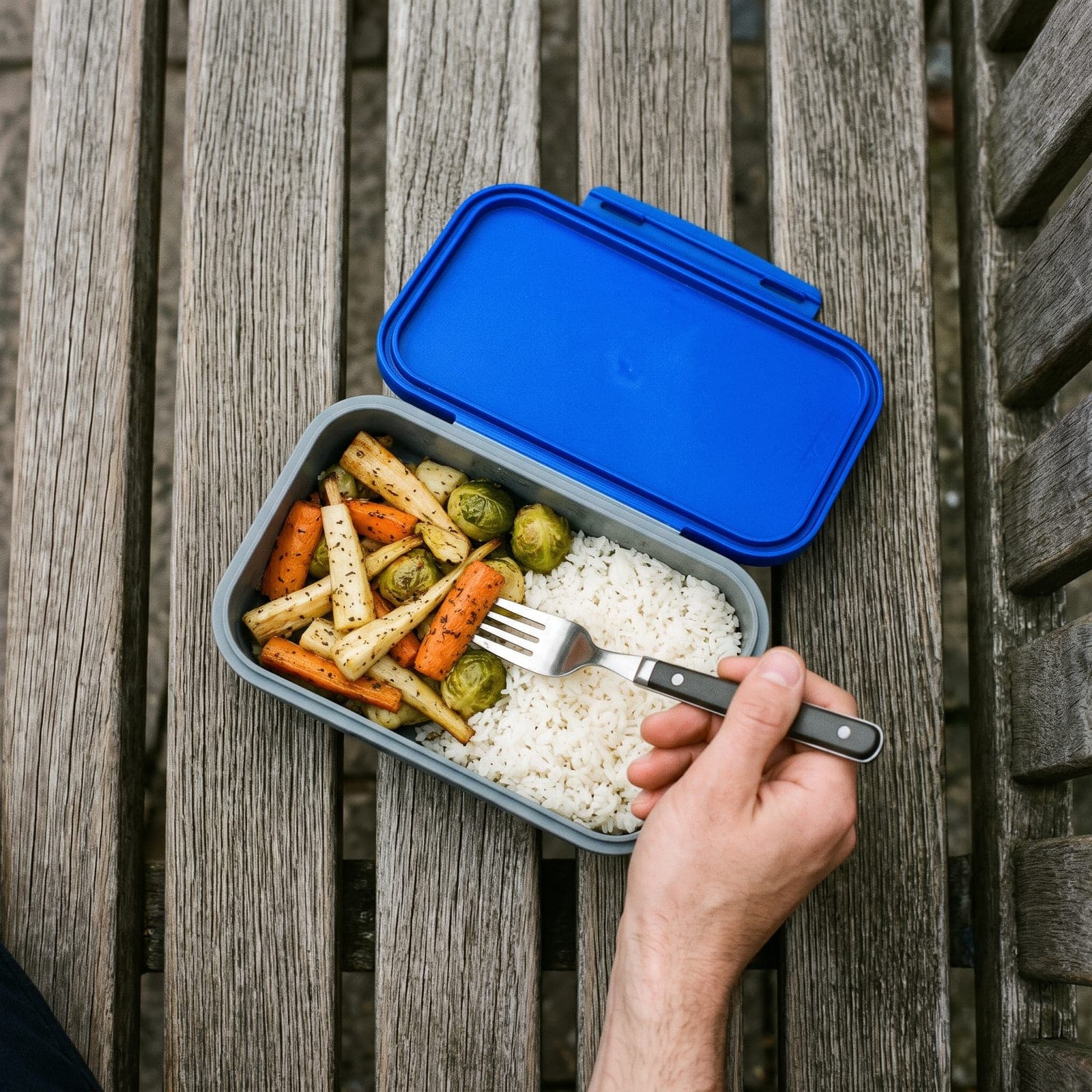 Boîte à déjeuner grise au couvercle bleu ouverte sur un banc, riz et légumes rôtis, une main tient une fourchette