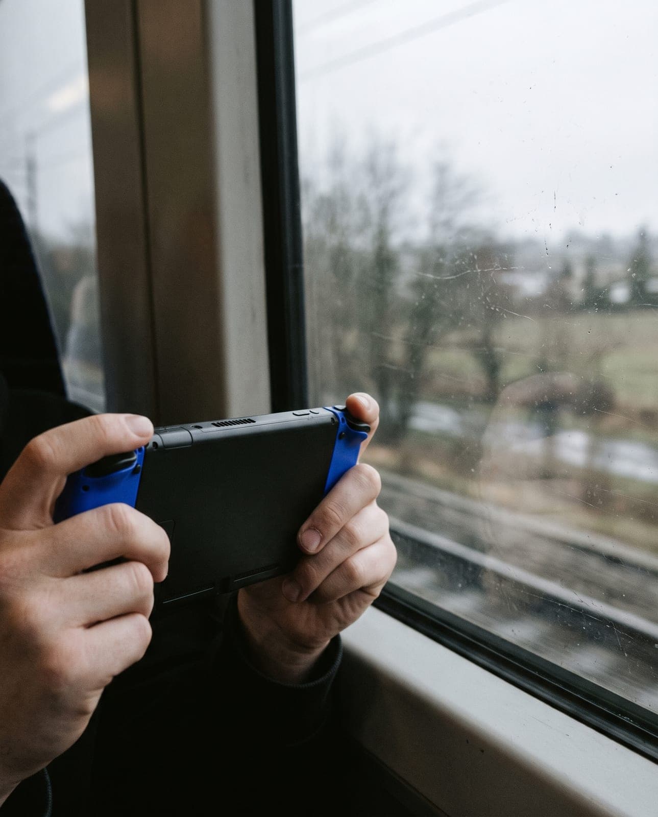 Console portable noire à l'écran éteint tenue à deux mains devant la fenêtre d'un train, capuchons de joysticks bleus