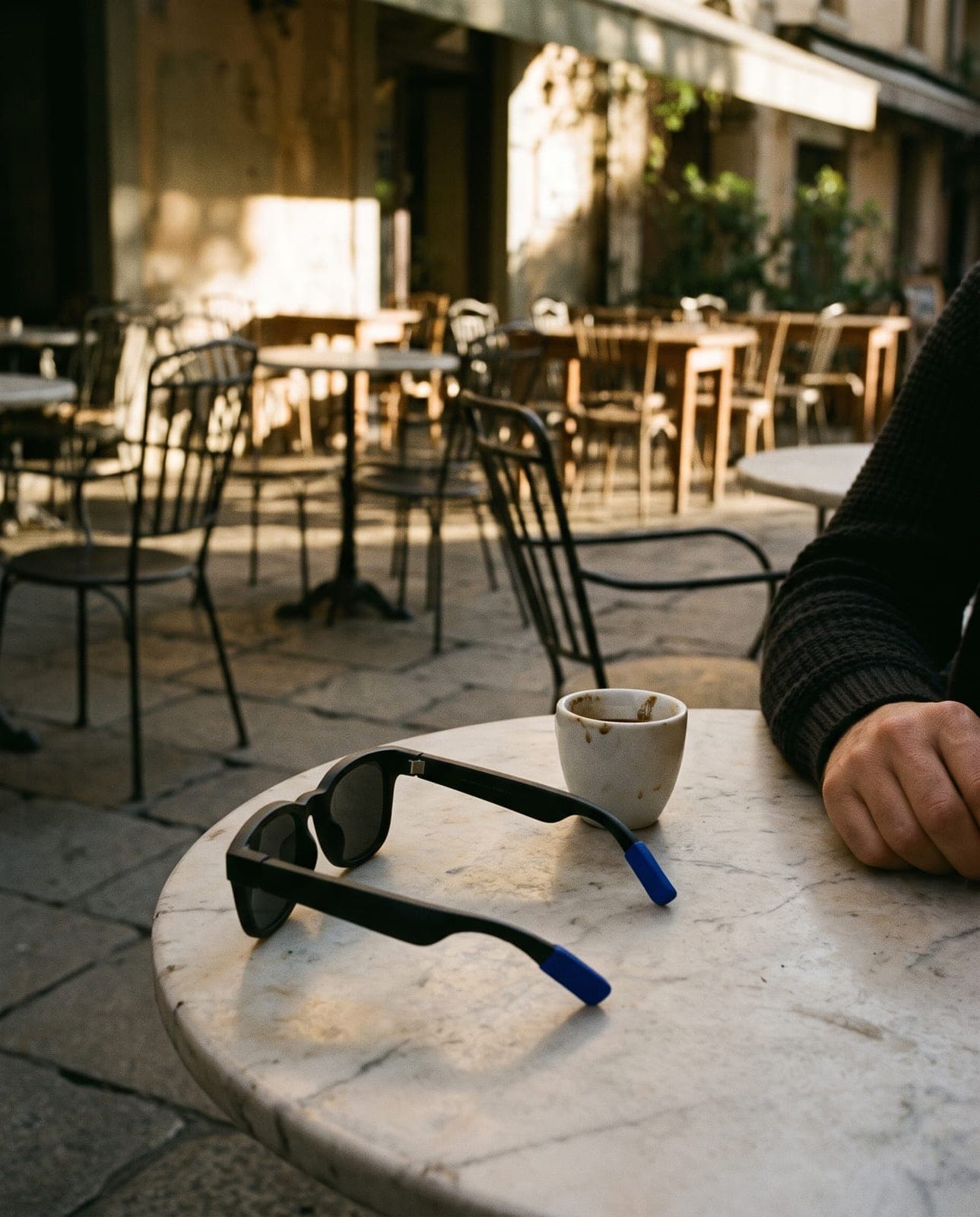 Black smart glasses with blue temple tips folded on a marble café table beside an espresso, a resting hand