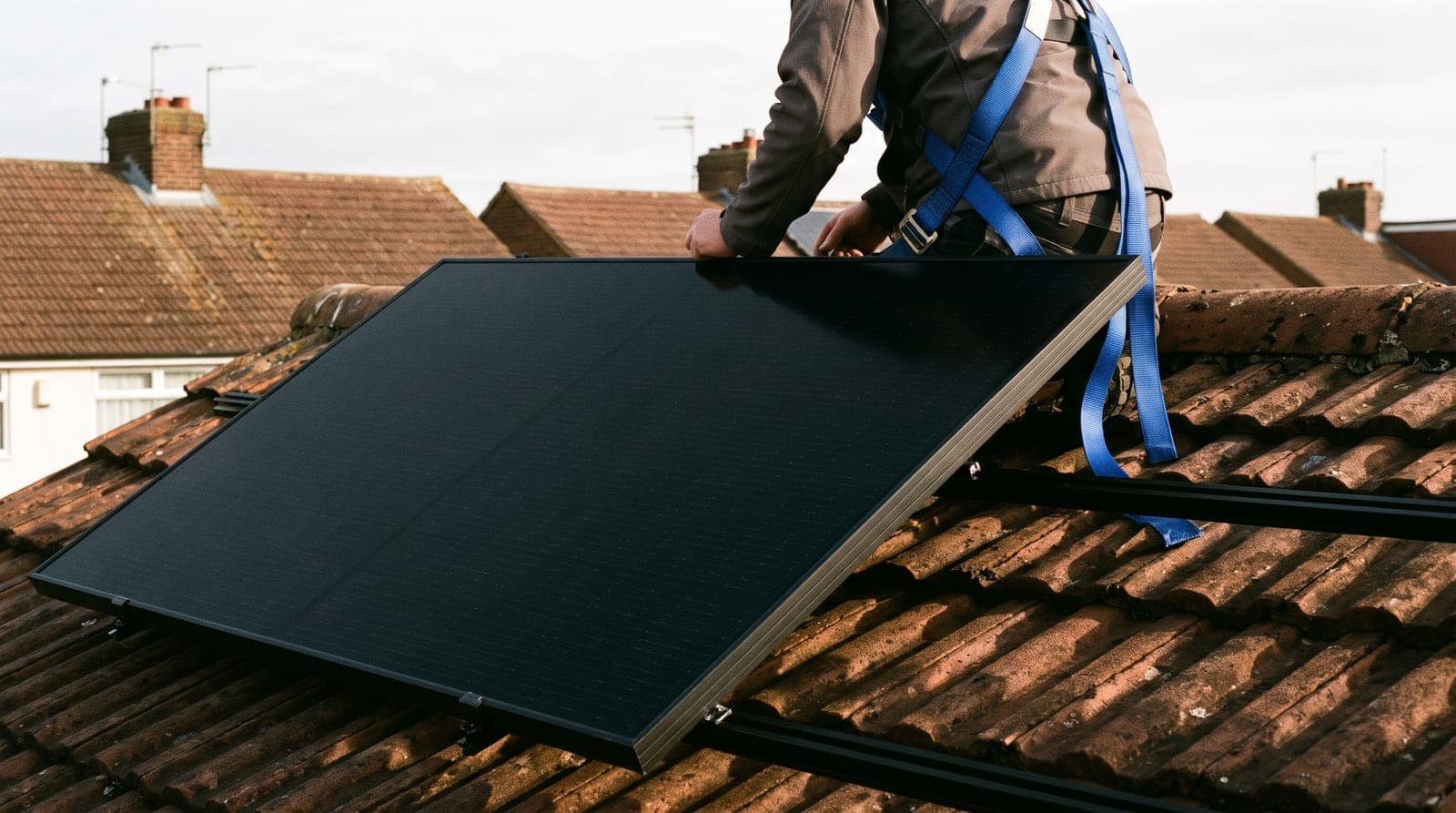Panneau solaire noir posé sur son rail sur un toit de tuiles, l'installateur accroupi de dos au harnais bleu