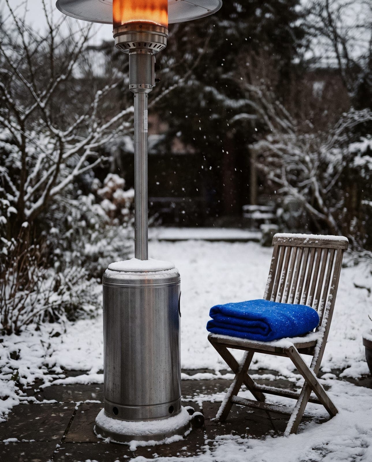 Patio heater glowing in a snowy garden at dusk, a blue blanket folded on a snow-covered chair