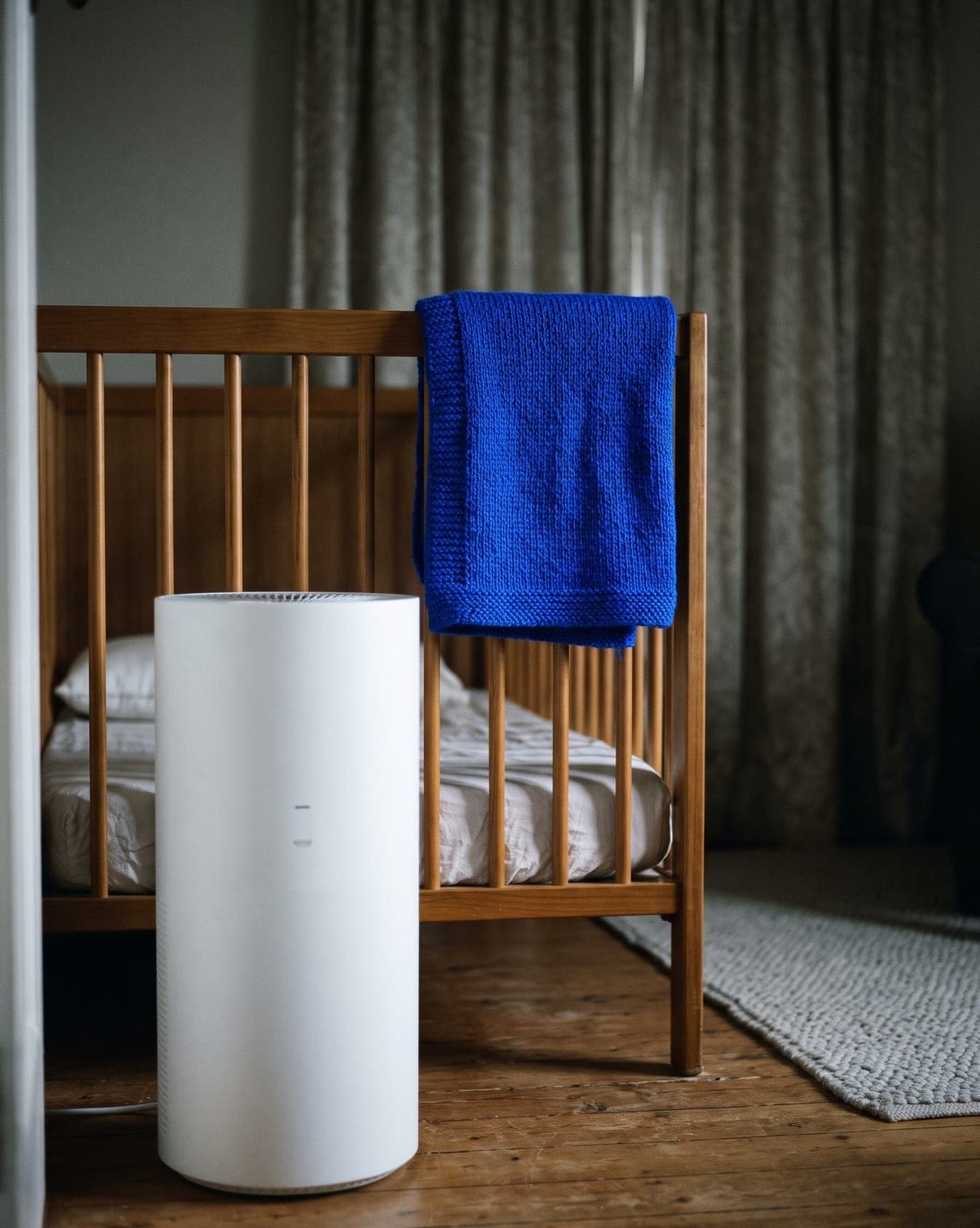 Purificateur d'air blanc au pied d'un lit à barreaux en bois, une couverture tricotée bleue sur la barrière