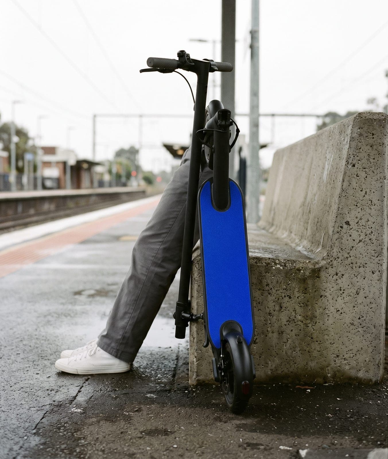 Folded electric scooter with blue grip tape leaning on a station platform bench, white trainers beside it